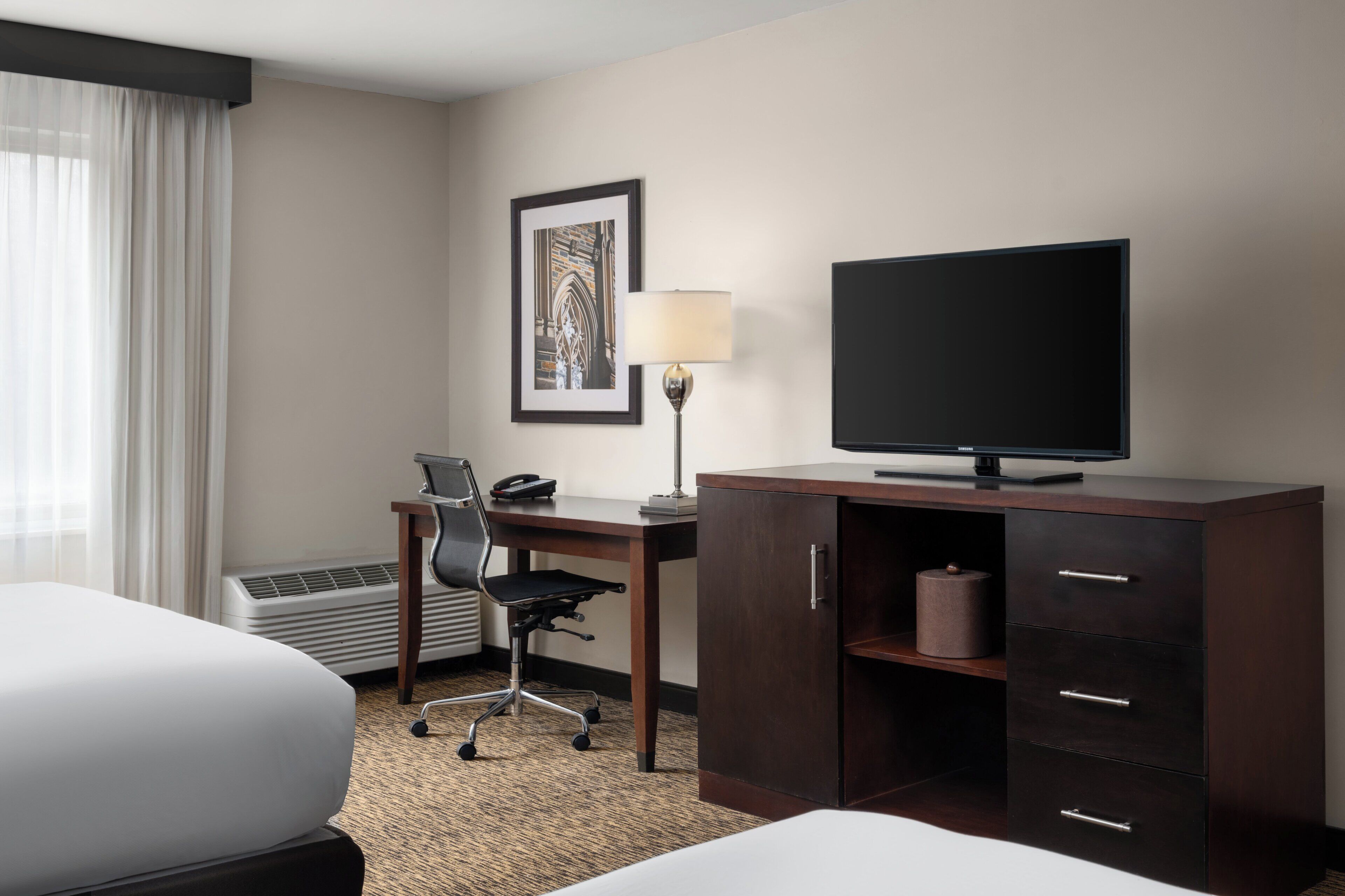A hotel room with a bed, desk, chair, television, lamp, and a framed picture on the wall.