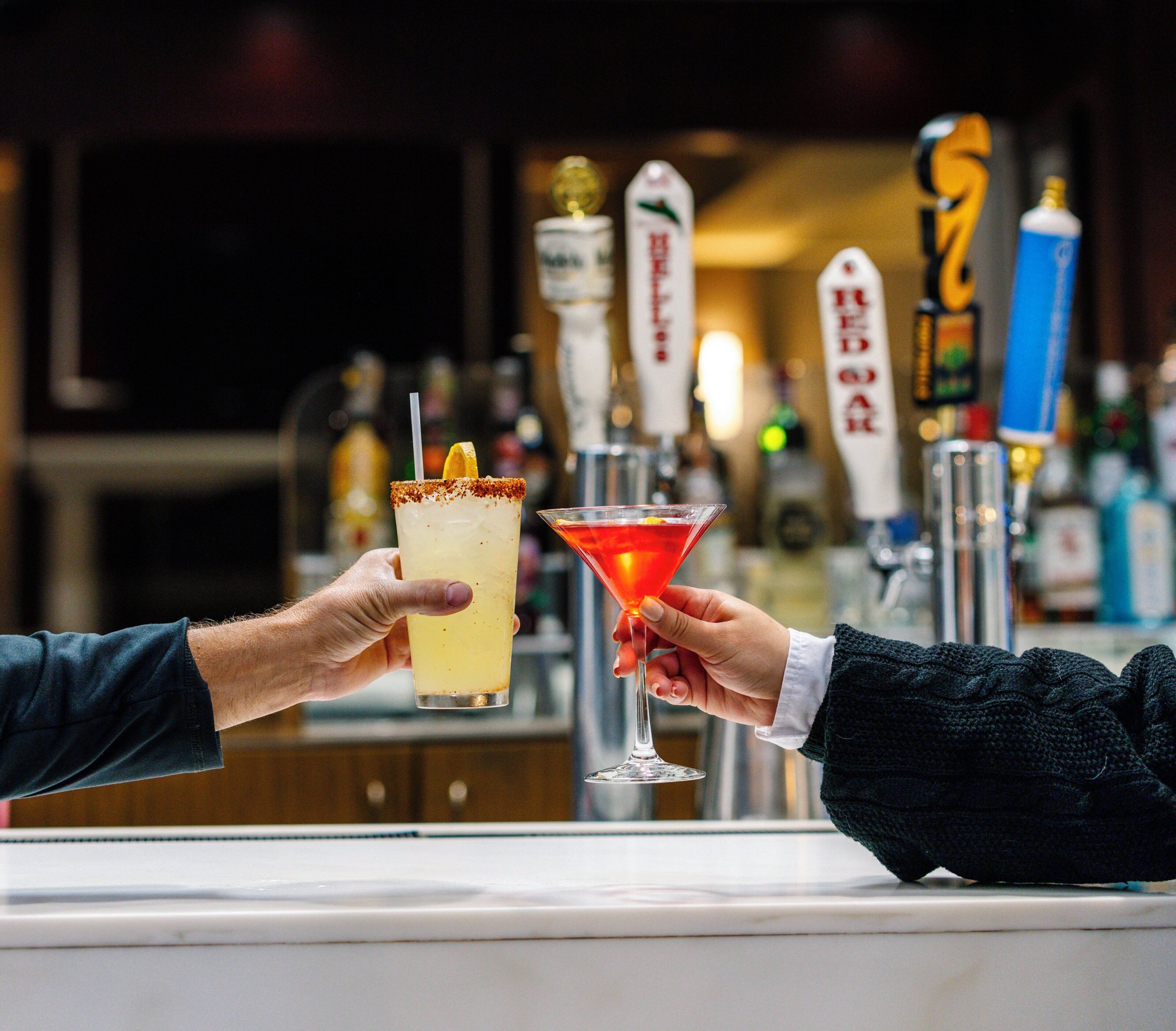 Two hands holding a margarita and a cocktail, with a bar and taps in the background.