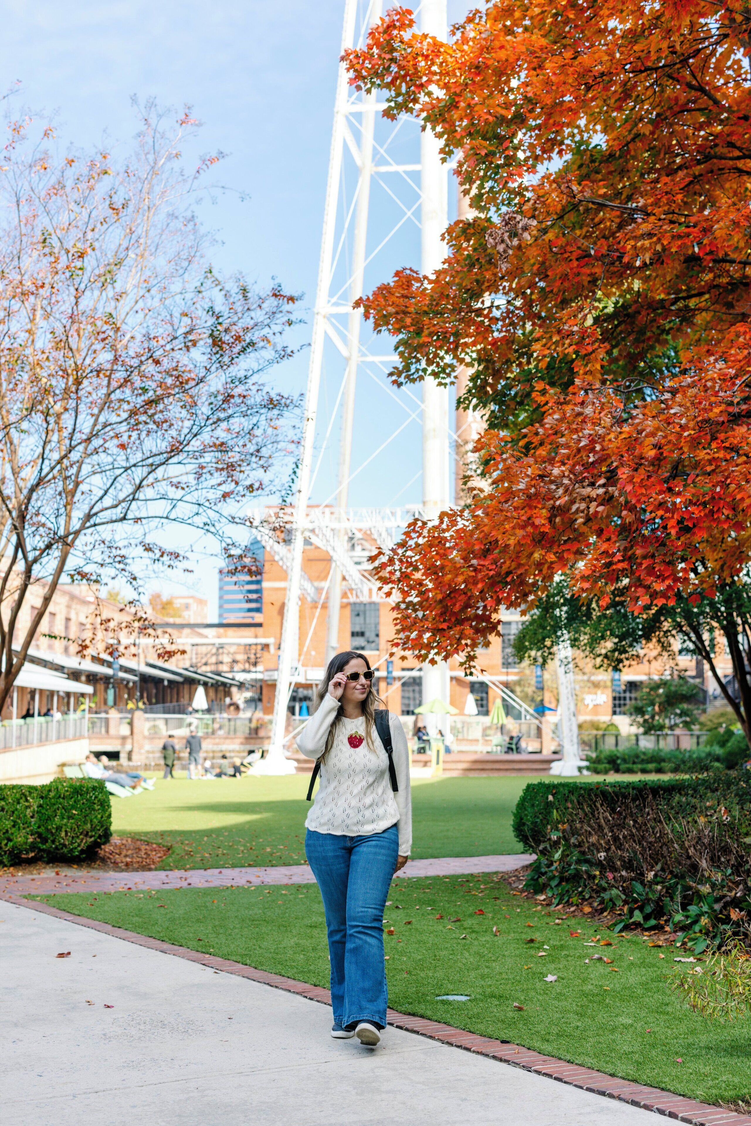 A woman walking on a pathway with autumn foliage and a white structure in the background.