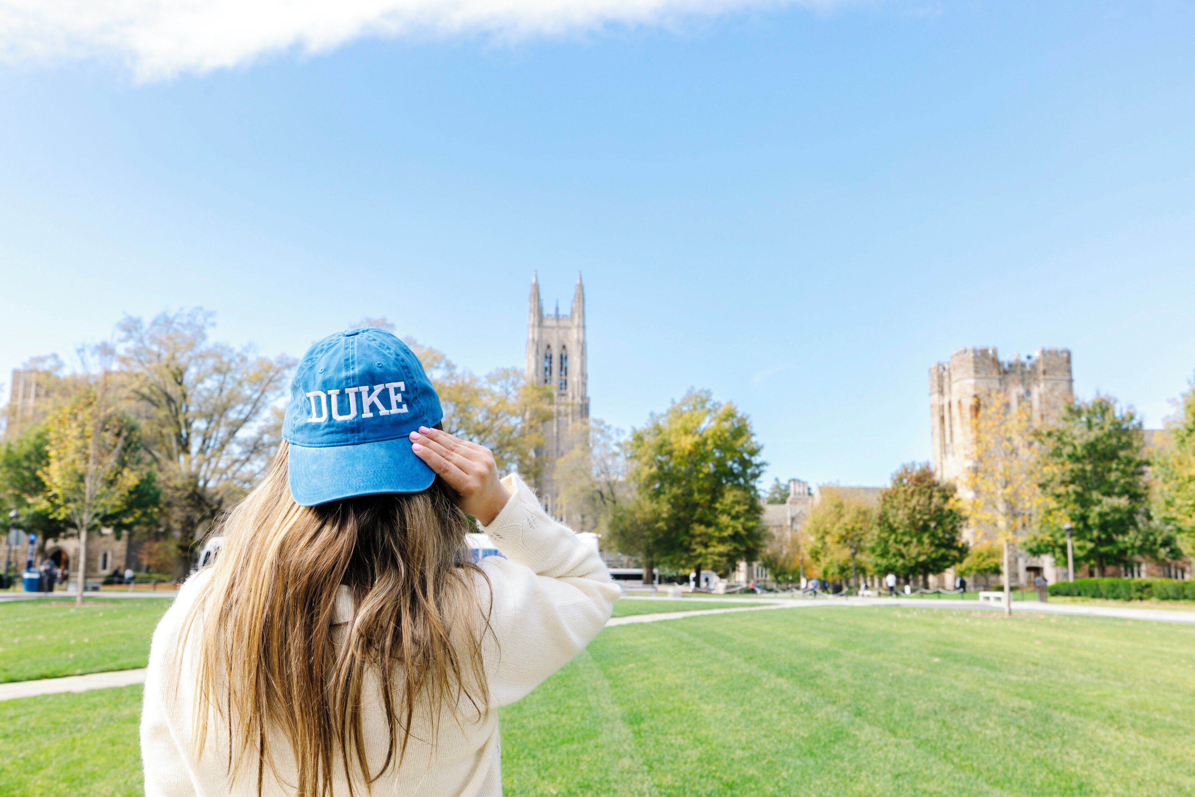 Una persona con una gorra que muestra la palabra 'DUKE', de pie en una zona cubierta de césped, con cielo despejado y edificios al fondo.