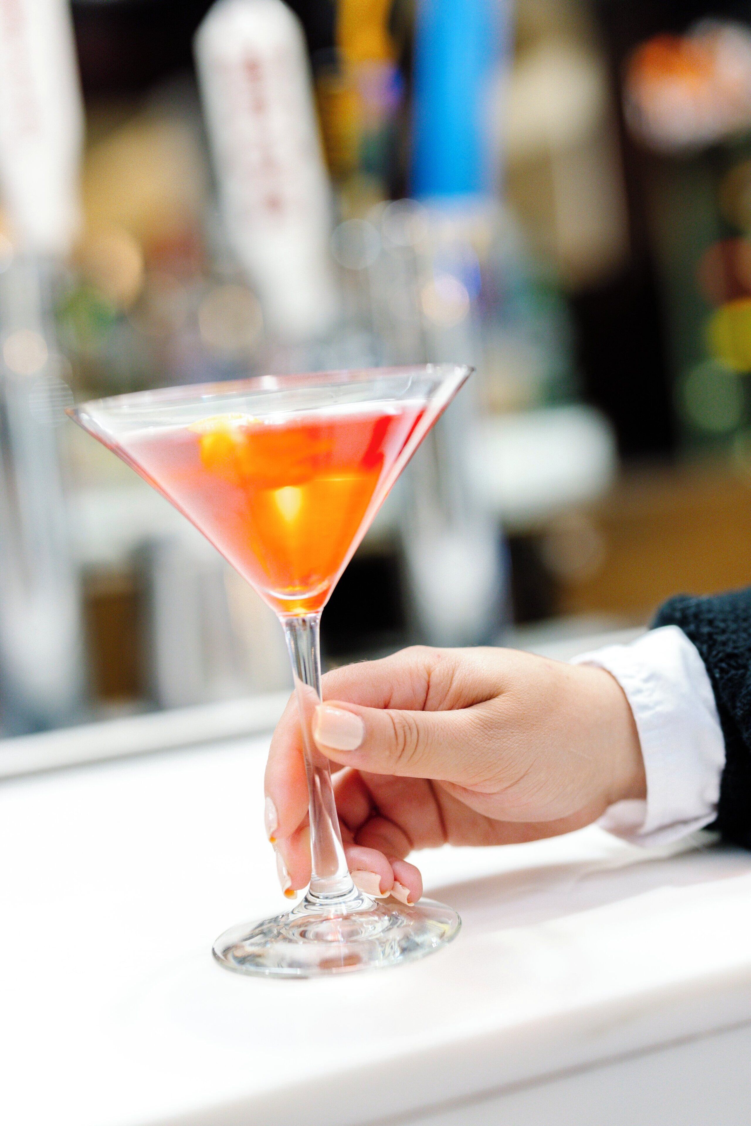 A hand holding a martini glass with a pink drink, set against a blurred background of a bar setting.