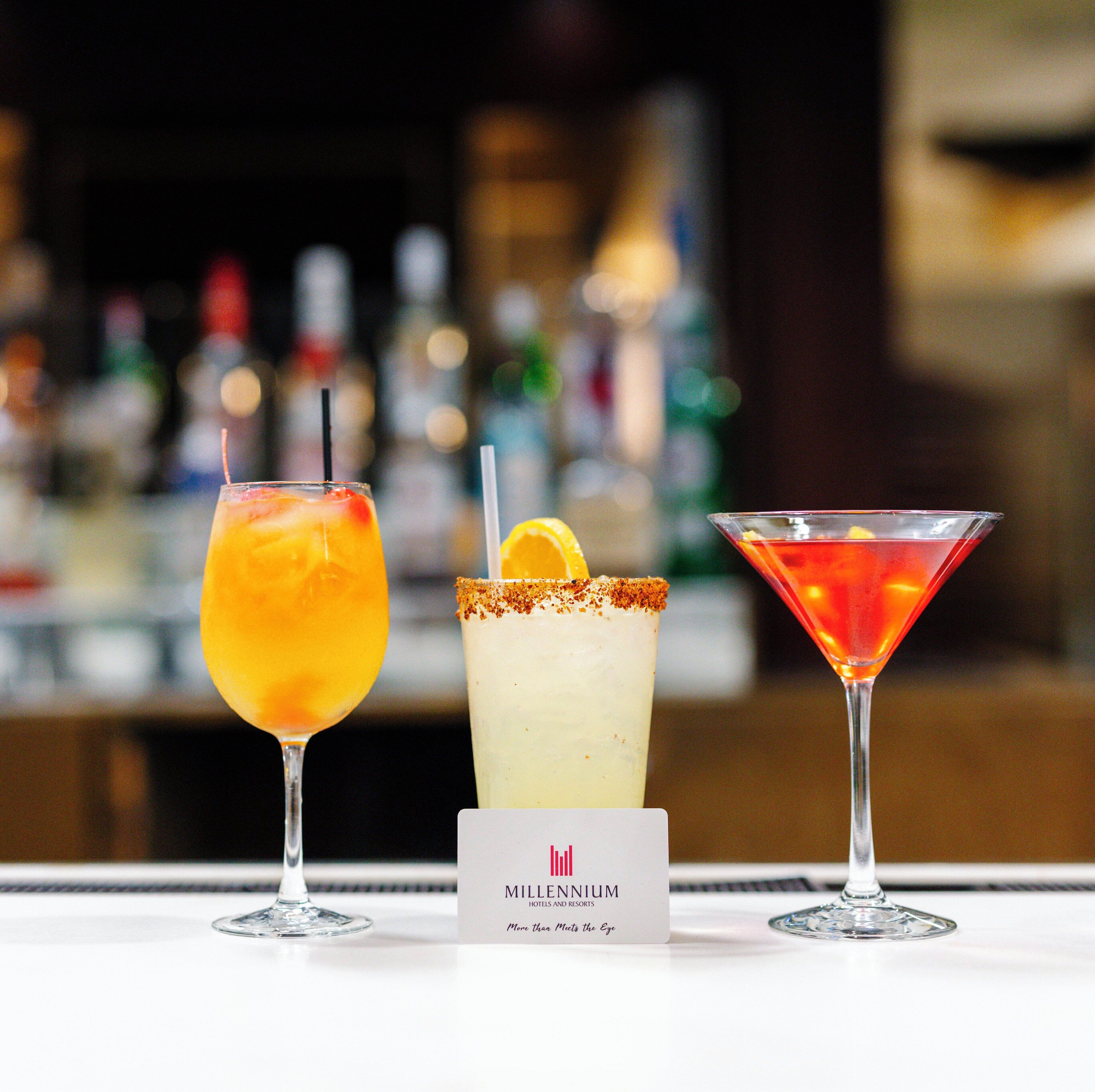 Three cocktails on a bar counter with a blurred background of bottles.