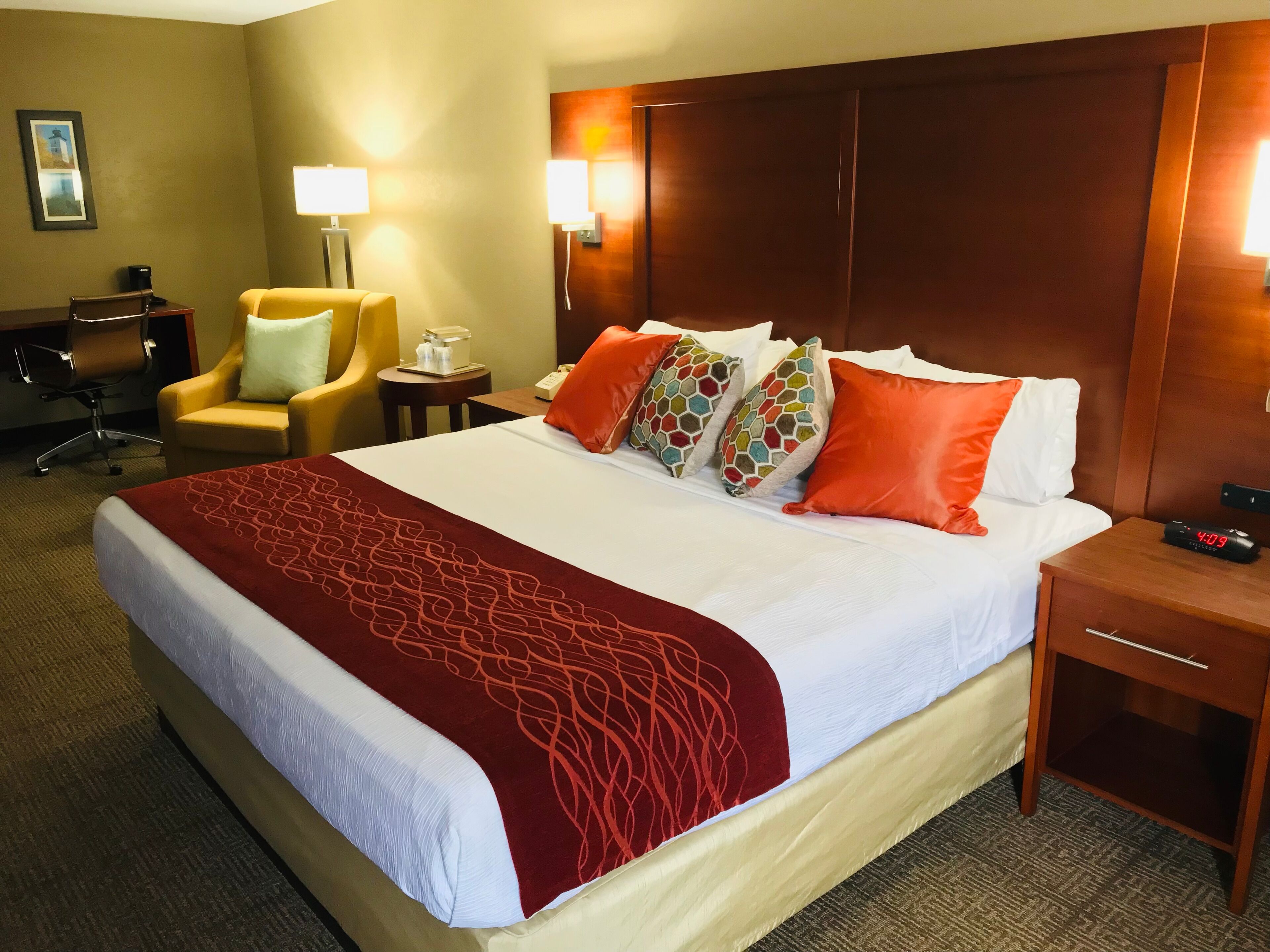 A hotel room with a large bed, a desk, a chair, a lamp, and a nightstand with a clock.