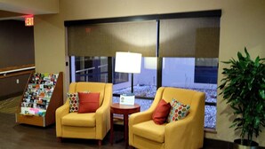 A lobby with two yellow armchairs, a small table with a lamp, a book display, and a large window with blinds. - Comfort Inn, Erie - Near Presque Isle (Erie)
