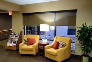 A lobby with two yellow armchairs, a small table with a lamp, a book display, and a large window with blinds.