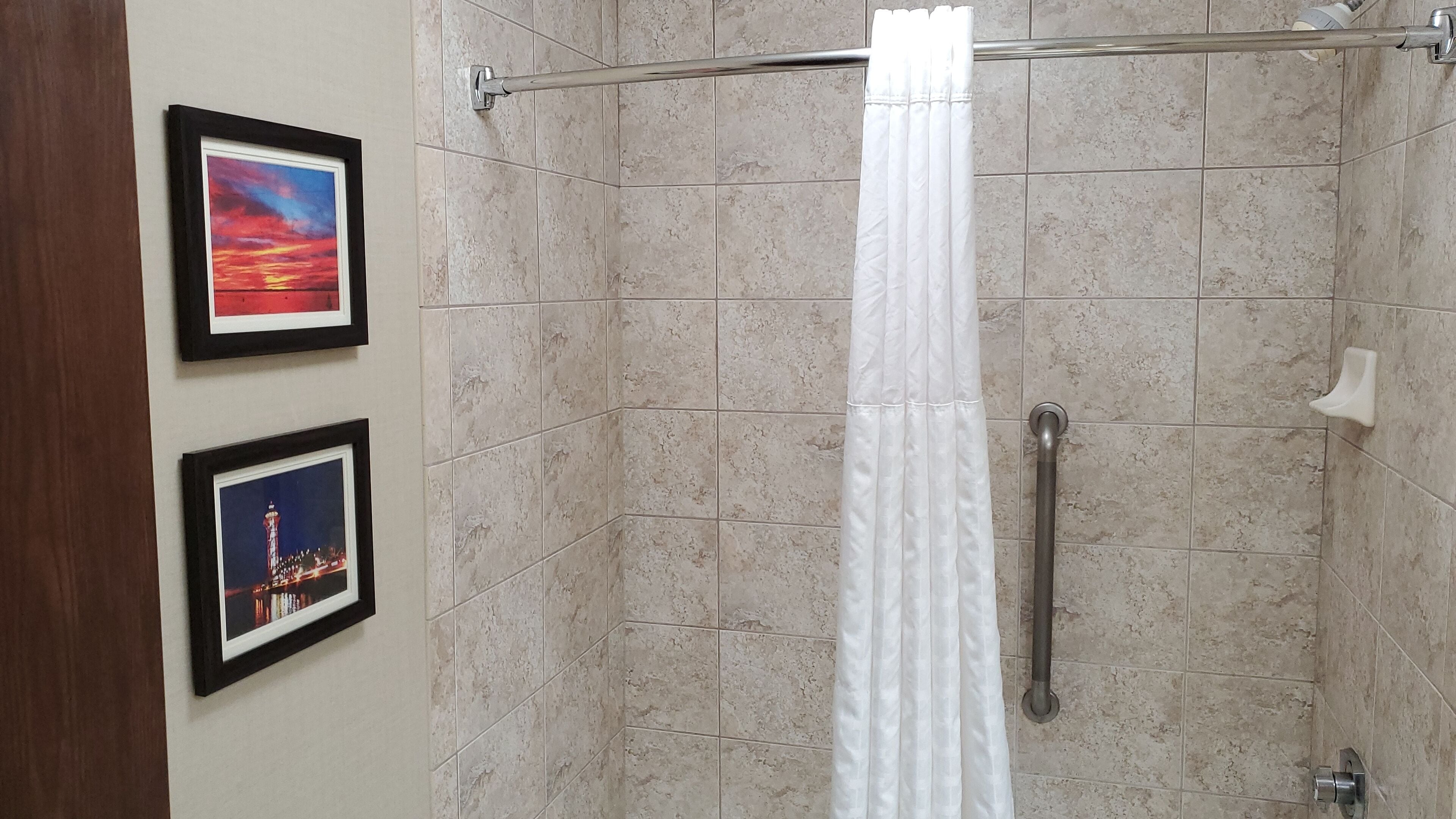 A shower area with beige tiles, a white shower curtain, and two framed pictures on the wall.