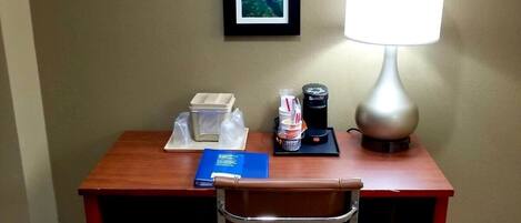 A hotel room with a wooden desk, a chair, a framed picture on the wall, a lamp, and a fire alarm.