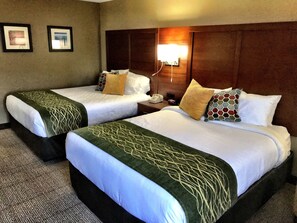 A hotel room with two beds, each with a green patterned bedspread, white pillows, and a wooden headboard. There is a nightstand with a phone and a lamp between the beds.