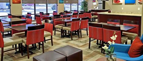 Un restaurante de hotel con varias mesas y sillas, una mesa de centro con un jarrón y vista al estacionamiento a través de grandes ventanales.