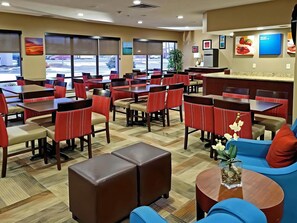 A hotel restaurant with multiple tables and chairs, a coffee table with a vase, and a view of the parking lot through large windows.