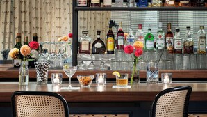 Una zona de bar con dos sillas de mimbre, diversas botellas de licores y una selección de vasos y flores sobre la barra.