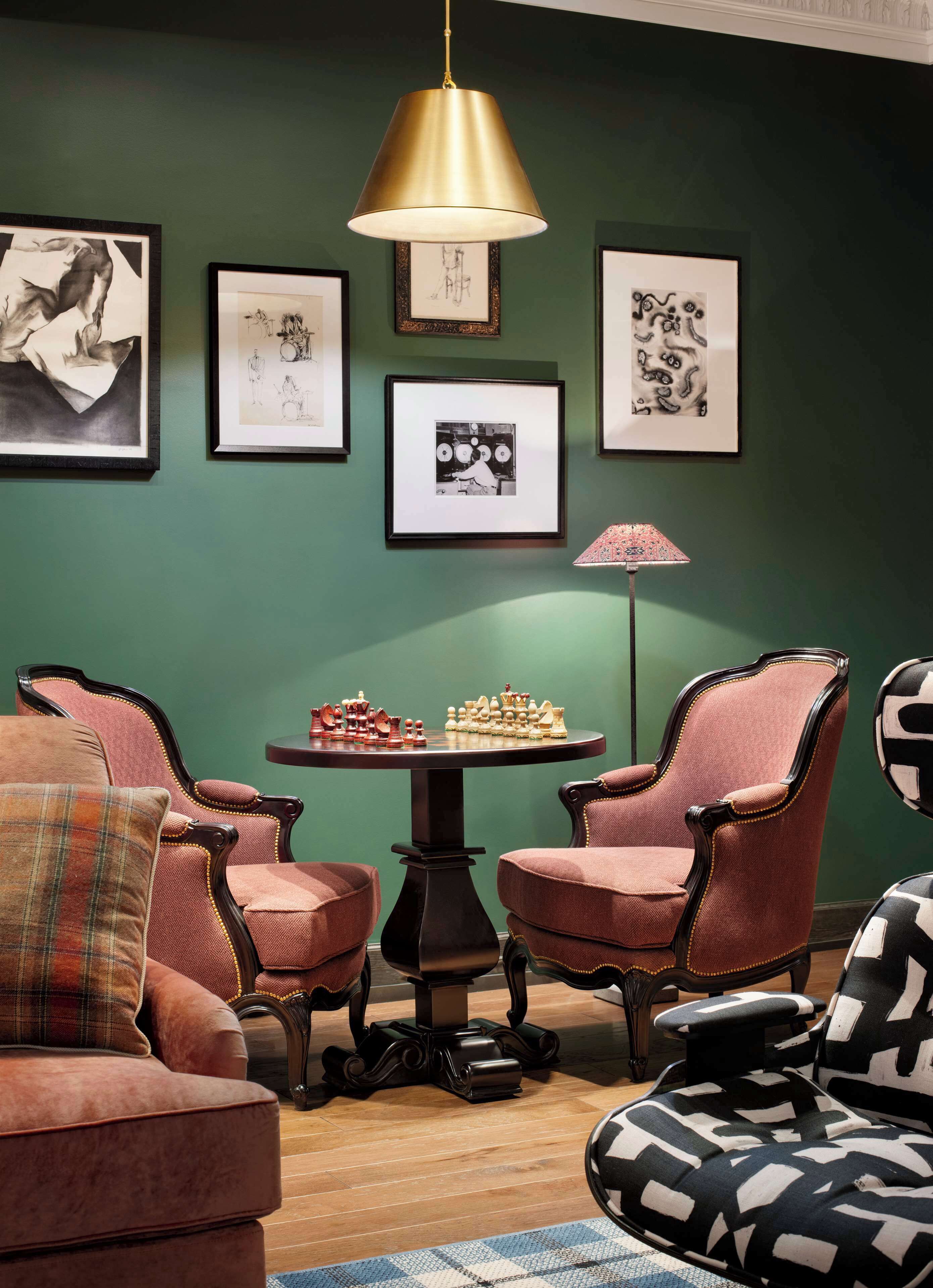 A room with green walls, a round table with chess set, two armchairs, a lamp, and framed artwork.