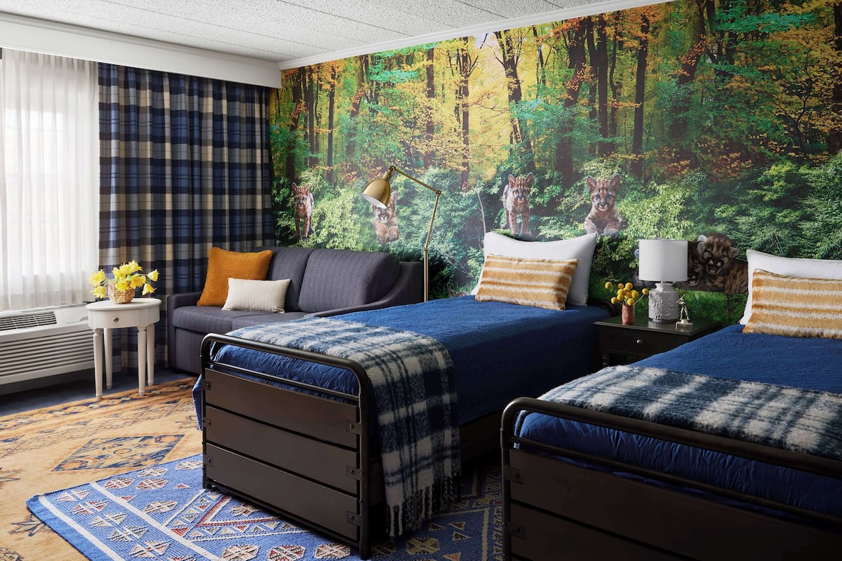 Ein Schlafzimmer mit zwei Betten, einem Schlafsofa, einer Lampe und einem Wandbild mit Waldmotiv.