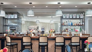 Zona de bar con diversas opciones de asientos, incluyendo taburetes y mesas redondas. Hay lámparas colgantes, una barra con diferentes botellas y vasos, y una máquina de café al fondo.