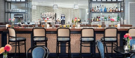 A bar area with multiple seating options, including bar stools and round tables. There are hanging pendant lights, a bar counter with various bottles and glasses, and a coffee machine in the background.