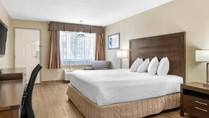 Ein Hotelzimmer mit einem großen Bett, einem Schreibtisch, einem Sessel, einem Fernseher, einem Fenster mit Vorhängen und einer Wandlampe.