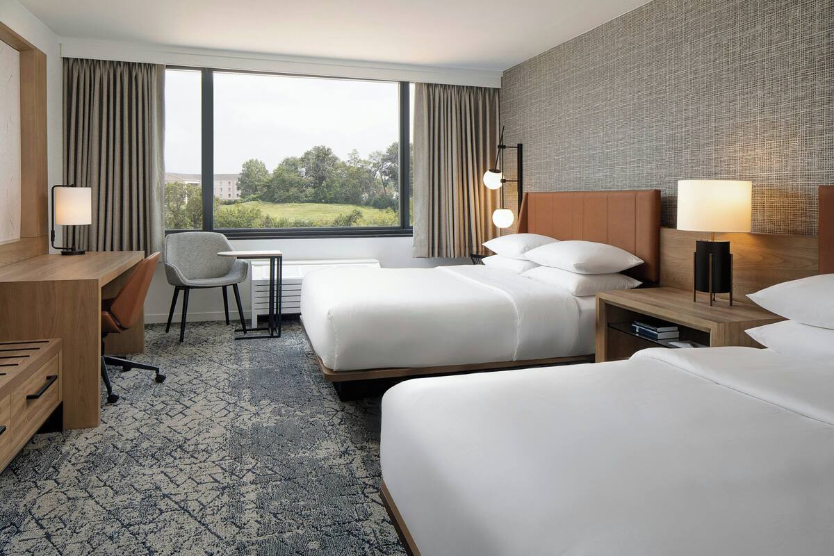 A hotel room with two beds, a desk, a chair, and a view of greenery outside.