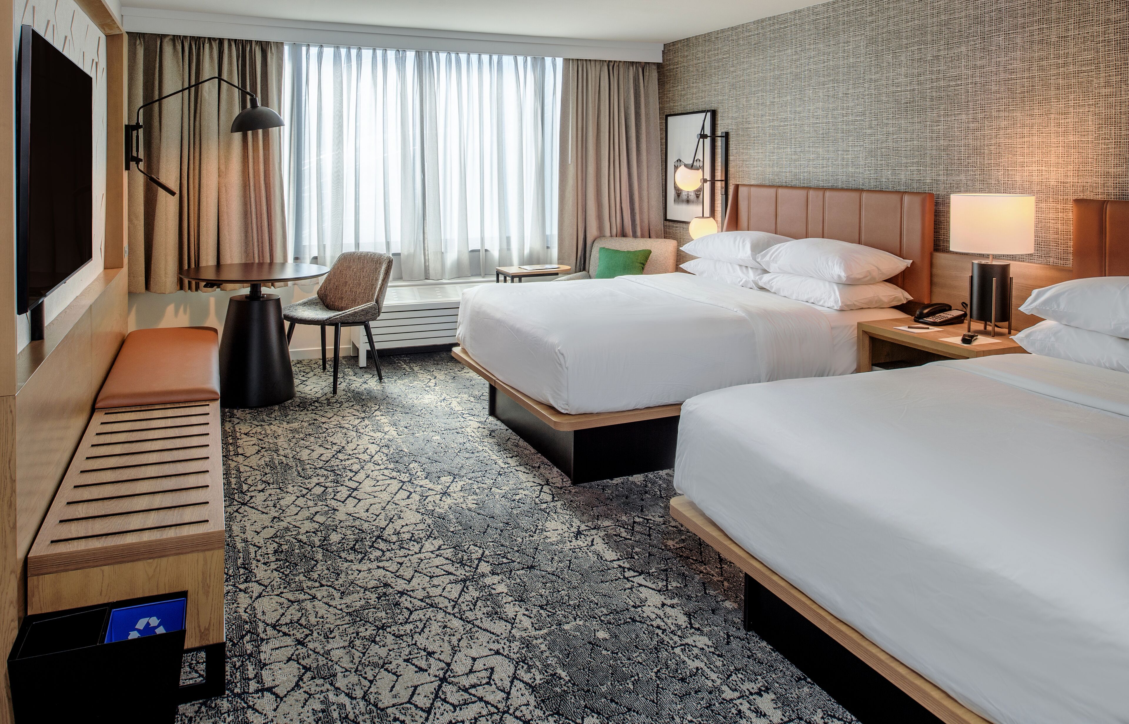 A hotel room with two beds, a desk, a chair, a TV, and a large window with curtains.