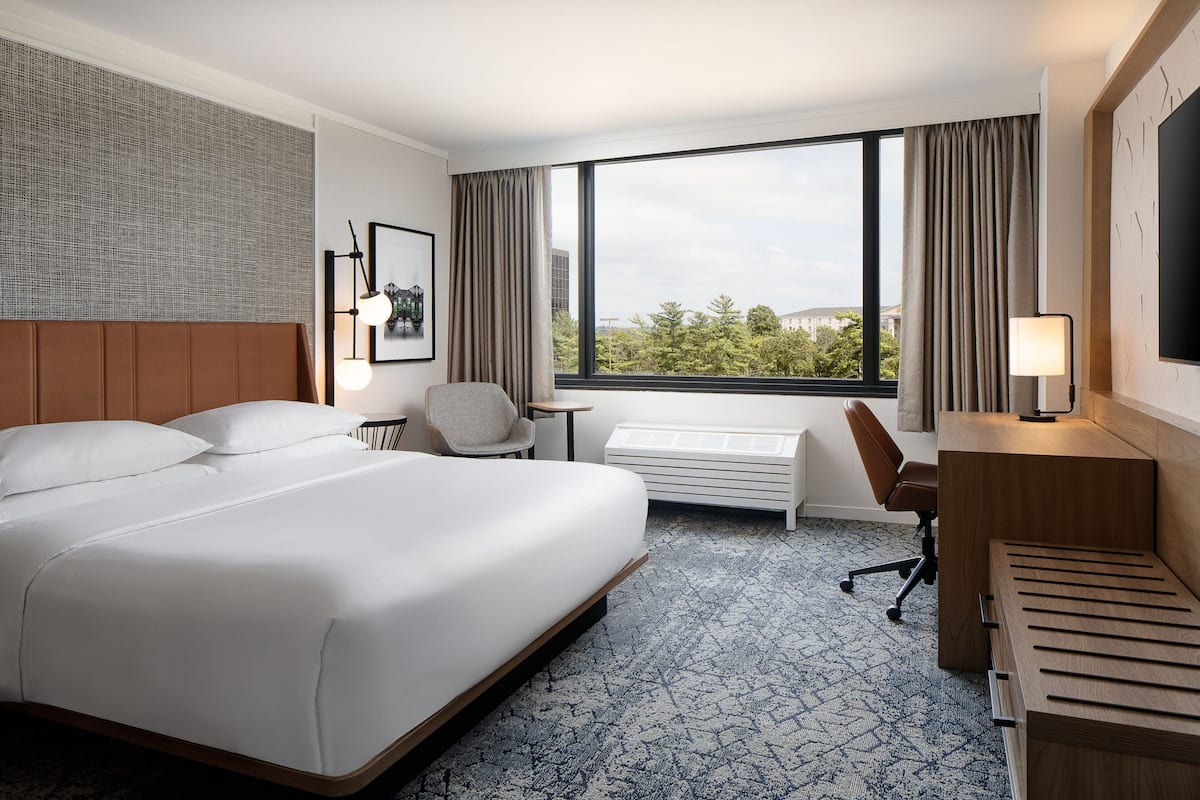 A hotel room with a large bed, a desk, a chair, a small table, a radiator, and a large window with curtains.