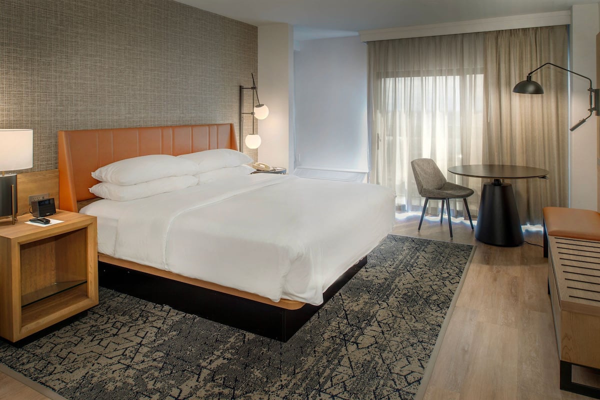 A hotel room with a large bed, a bedside table with a lamp, a chair, a small table, and a view of the outdoors through curtains.