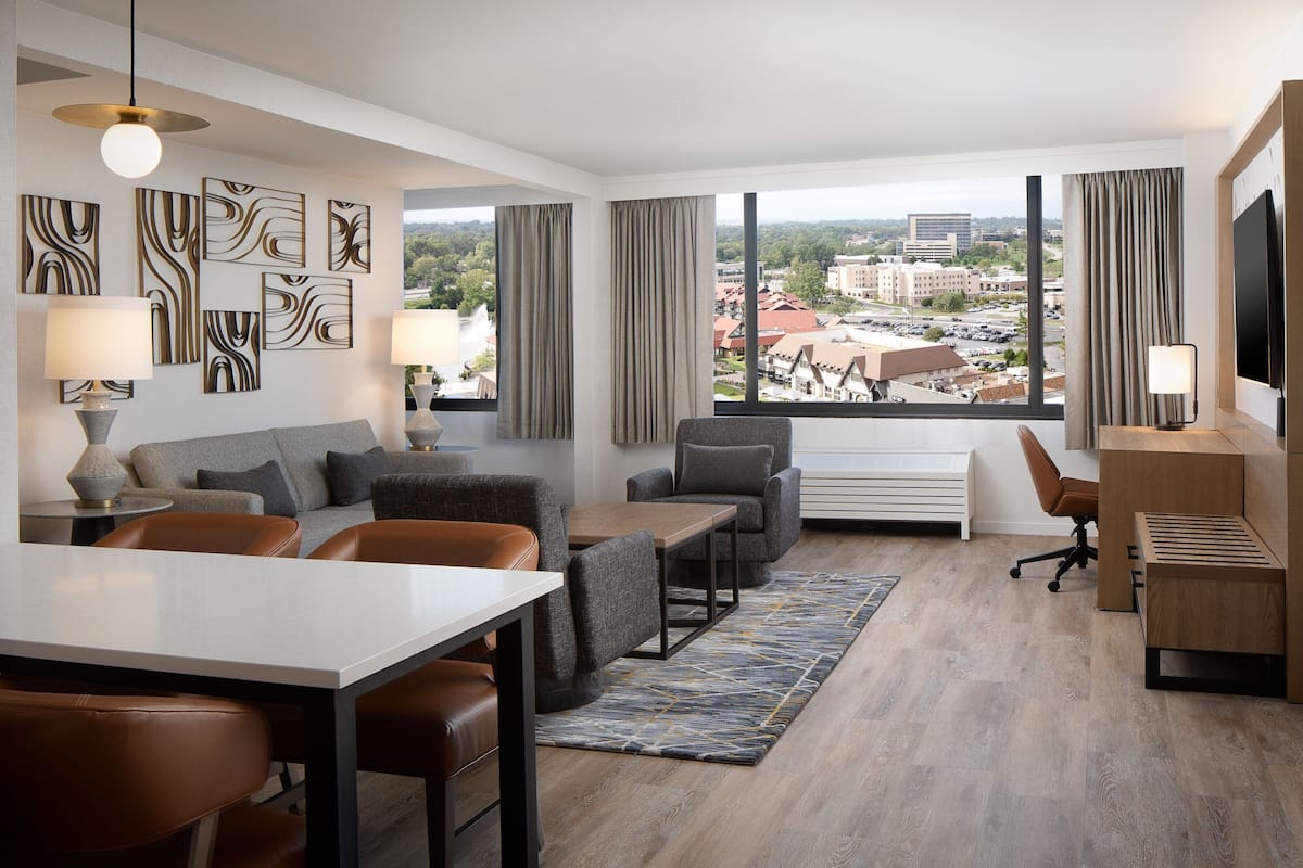 A modern hotel room with a dining area, a sofa set, a desk, and large windows offering a city view.