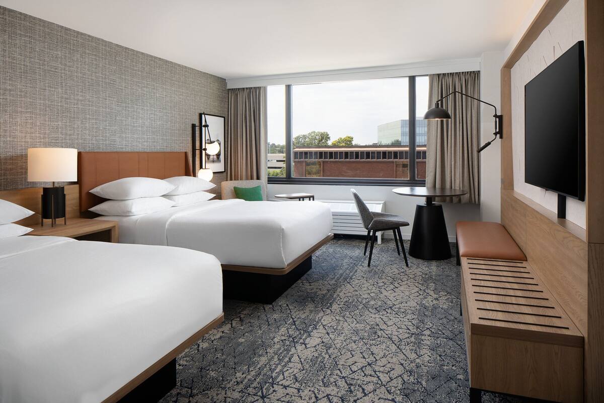A hotel room with two beds, a desk, a chair, a TV, and a large window with a view of buildings.