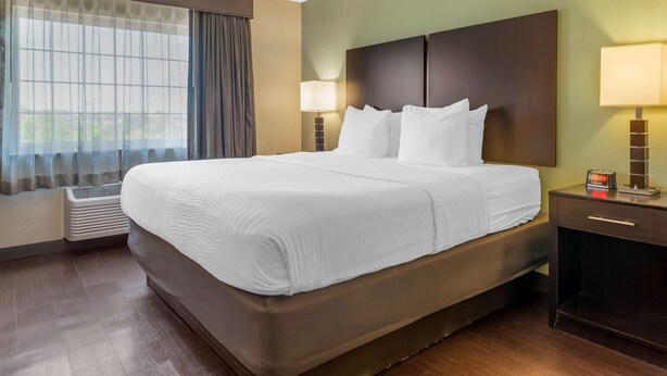 A hotel room with a large bed, a bedside table with a lamp, a window with curtains, and an air conditioning unit. - Best Western Hilliard Inn & Suites (Hilliard)