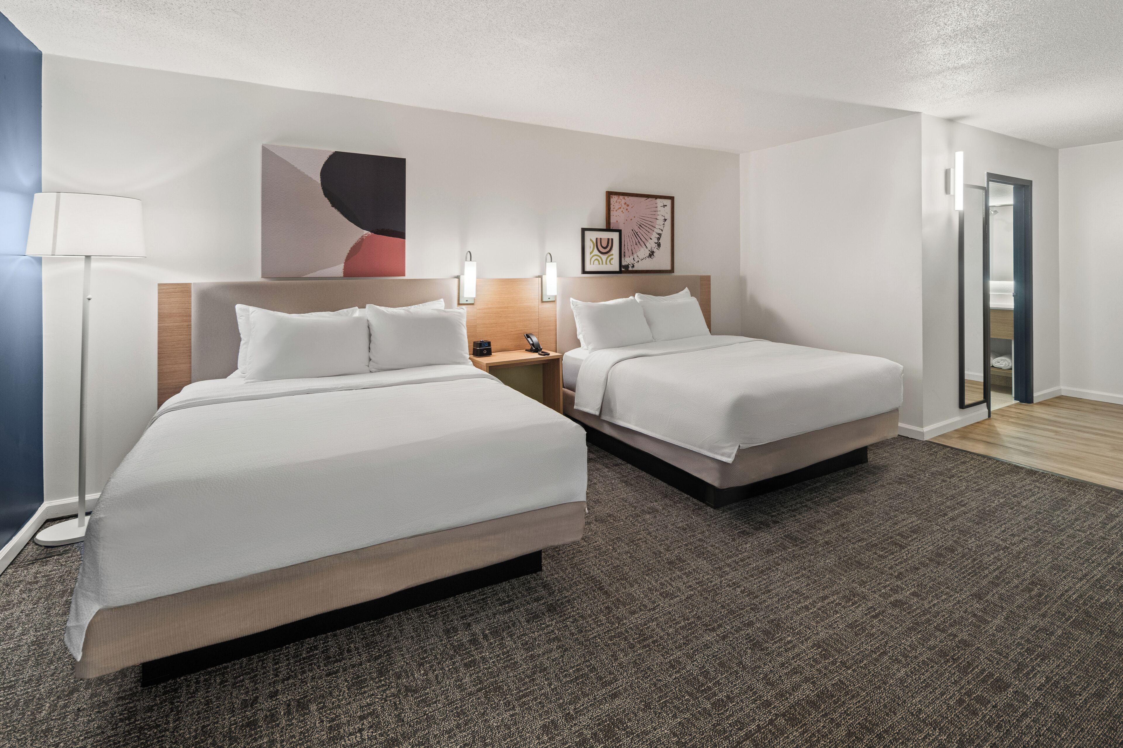 A hotel room with two beds, a bedside table with a lamp, and artwork on the walls.