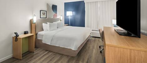 A hotel room with a bed, bedside tables, a desk, a flat-screen TV, and a lamp.