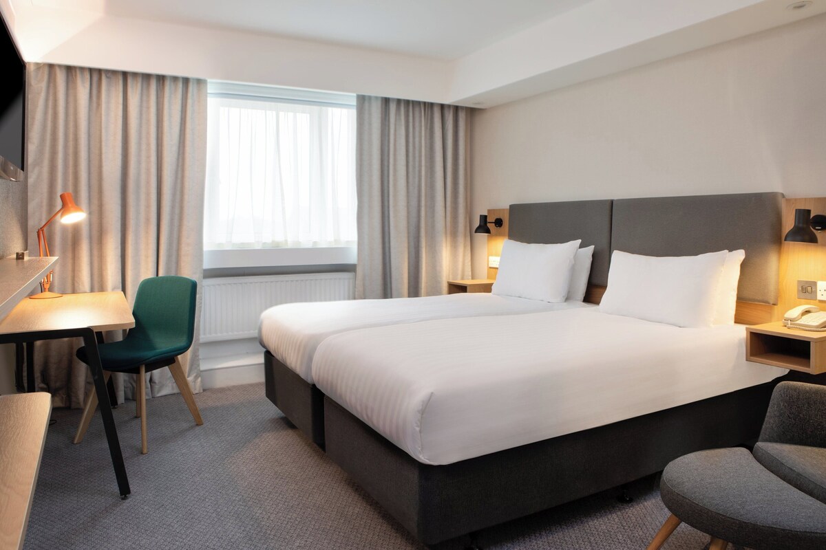 Une chambre d’hôtel avec un grand lit, un bureau, une chaise, une lampe et une fenêtre avec des rideaux.