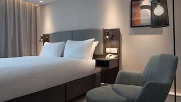 A modern hotel room with a large bed, a chair, a nightstand with a phone, and a wall-mounted lamp.