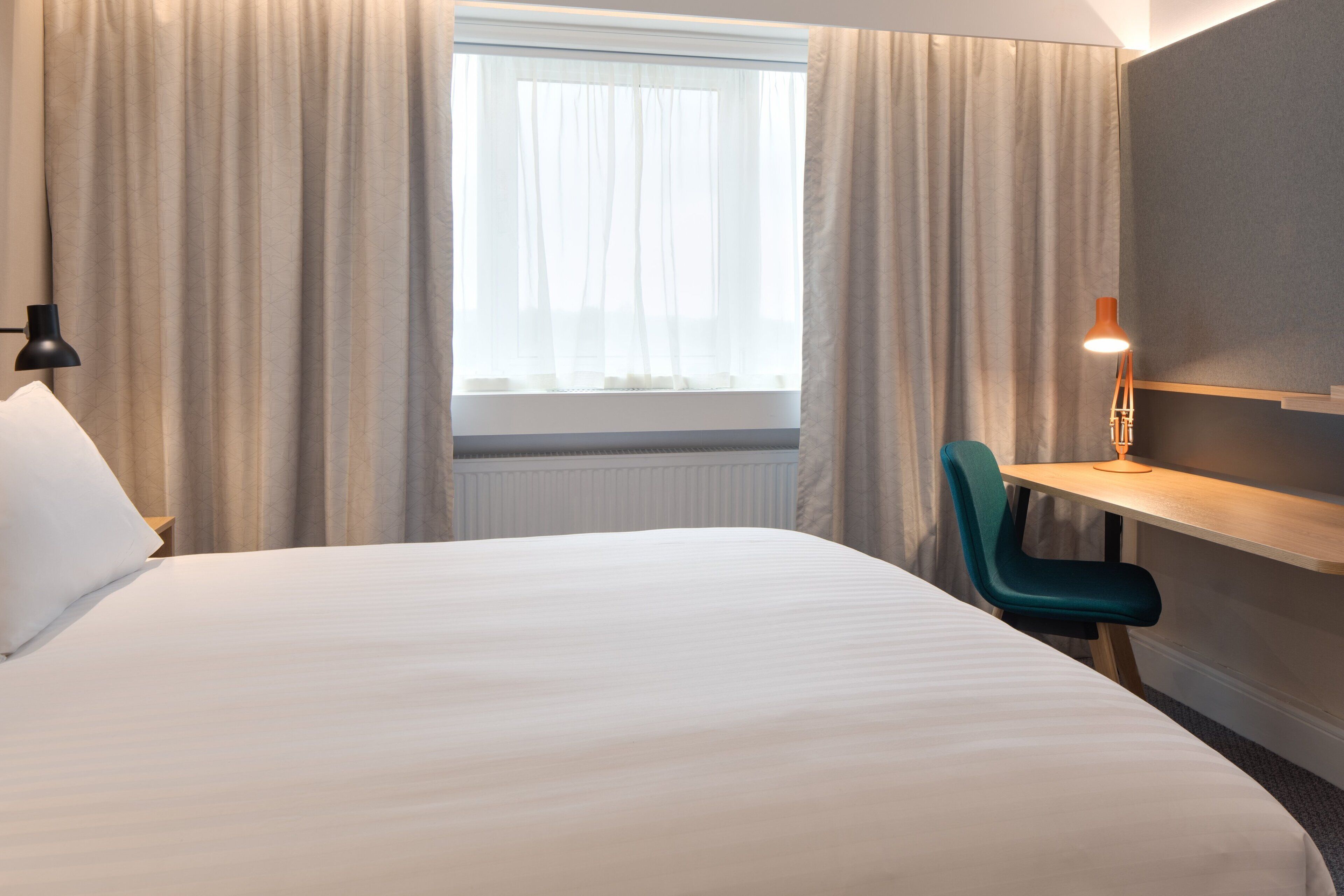 A hotel room with a bed, a desk, a chair, a lamp, and a window with curtains.