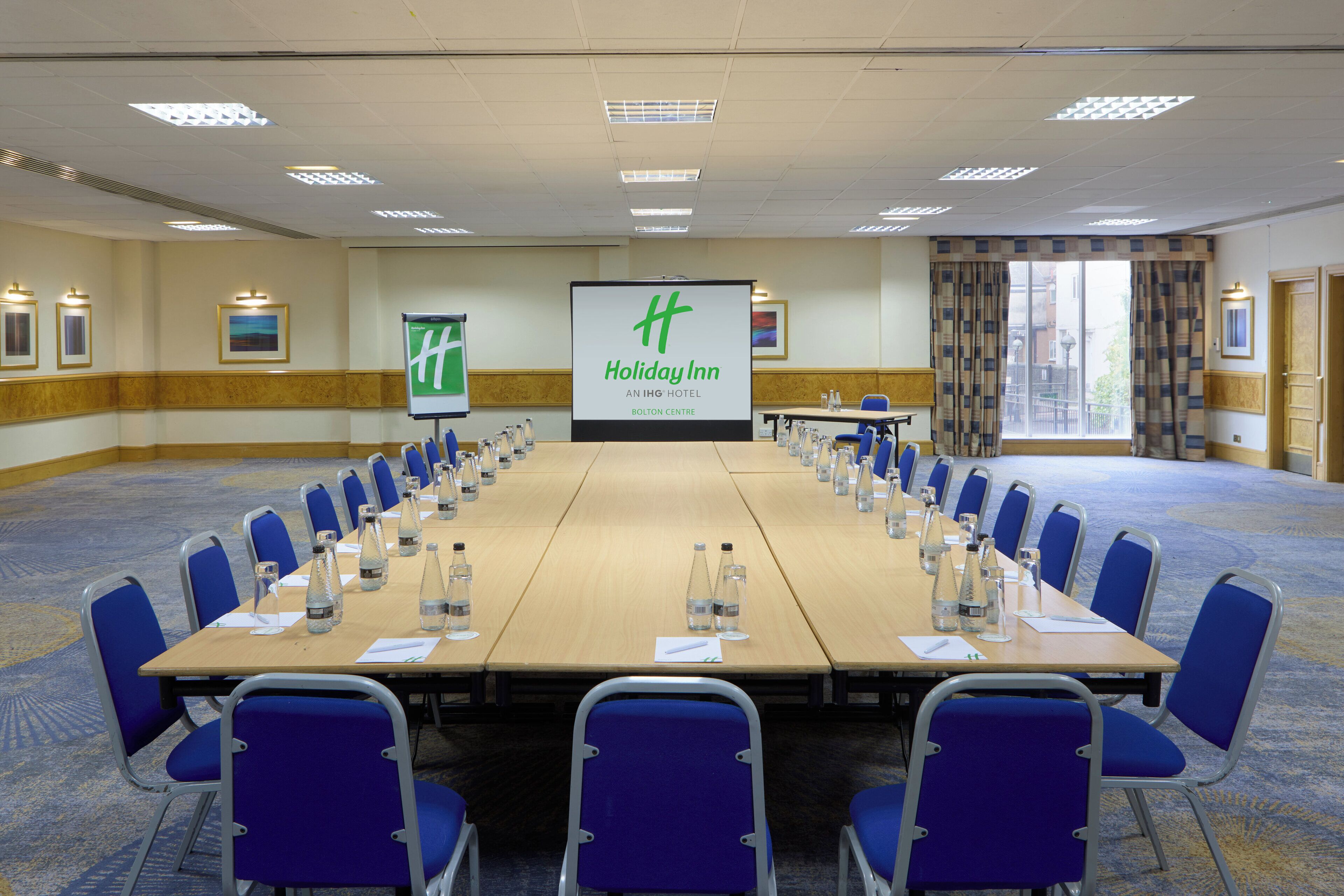 Ein Konferenzraum mit einem langen Tisch, blauen Stühlen und einem Bildschirm, der das Holiday Inn Logo zeigt.