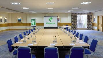 Ein Konferenzraum mit einem langen Tisch, blauen Stühlen und einem Bildschirm, der das Holiday Inn Logo zeigt.