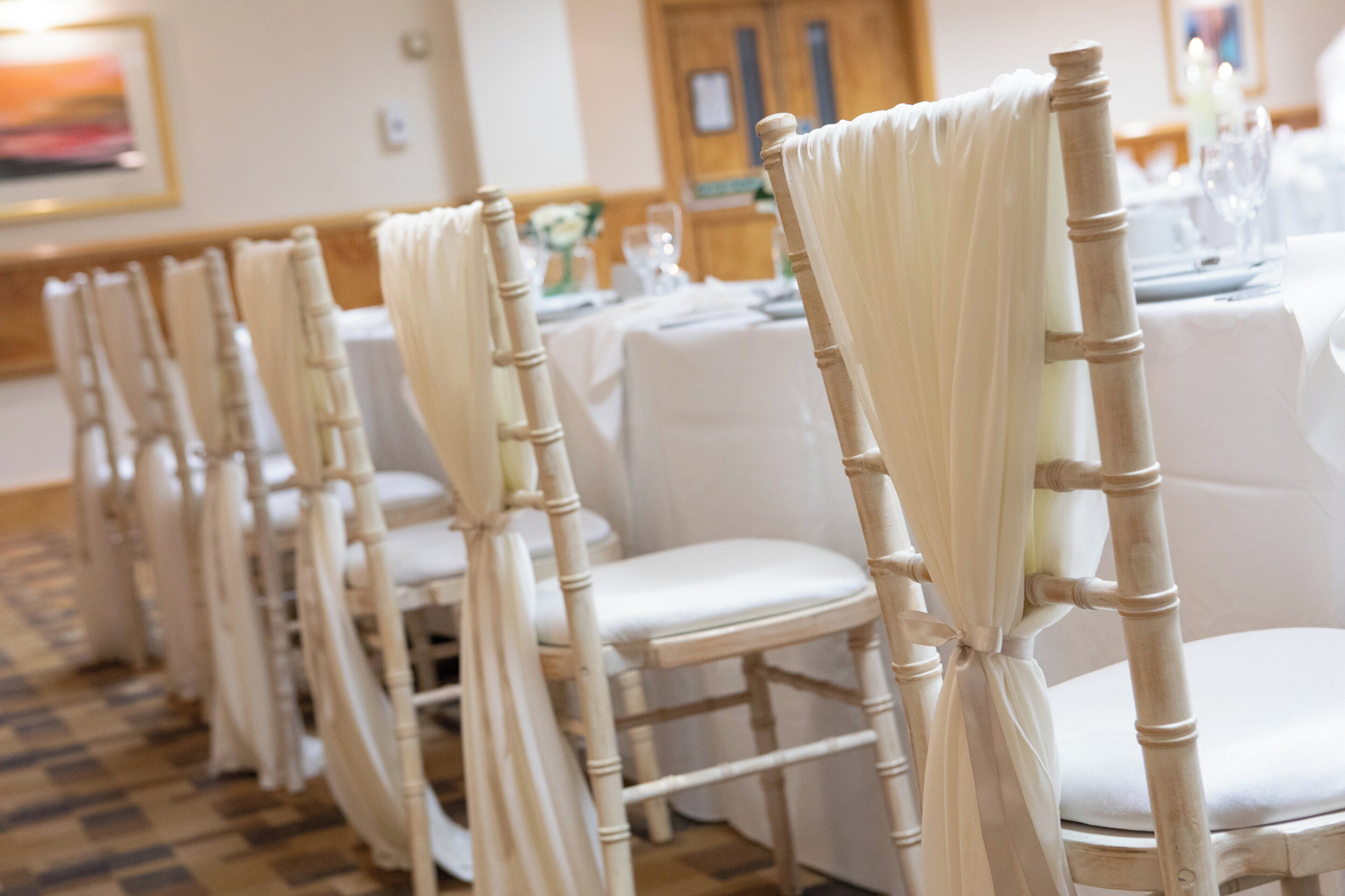 Rows of white chairs with cream-colored wooden frames and white fabric drapes, arranged in a banquet hall with a checkered floor pattern.