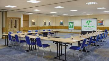 A conference room with multiple tables and chairs, a large screen displaying the Holiday Inn logo, and a carpeted floor.