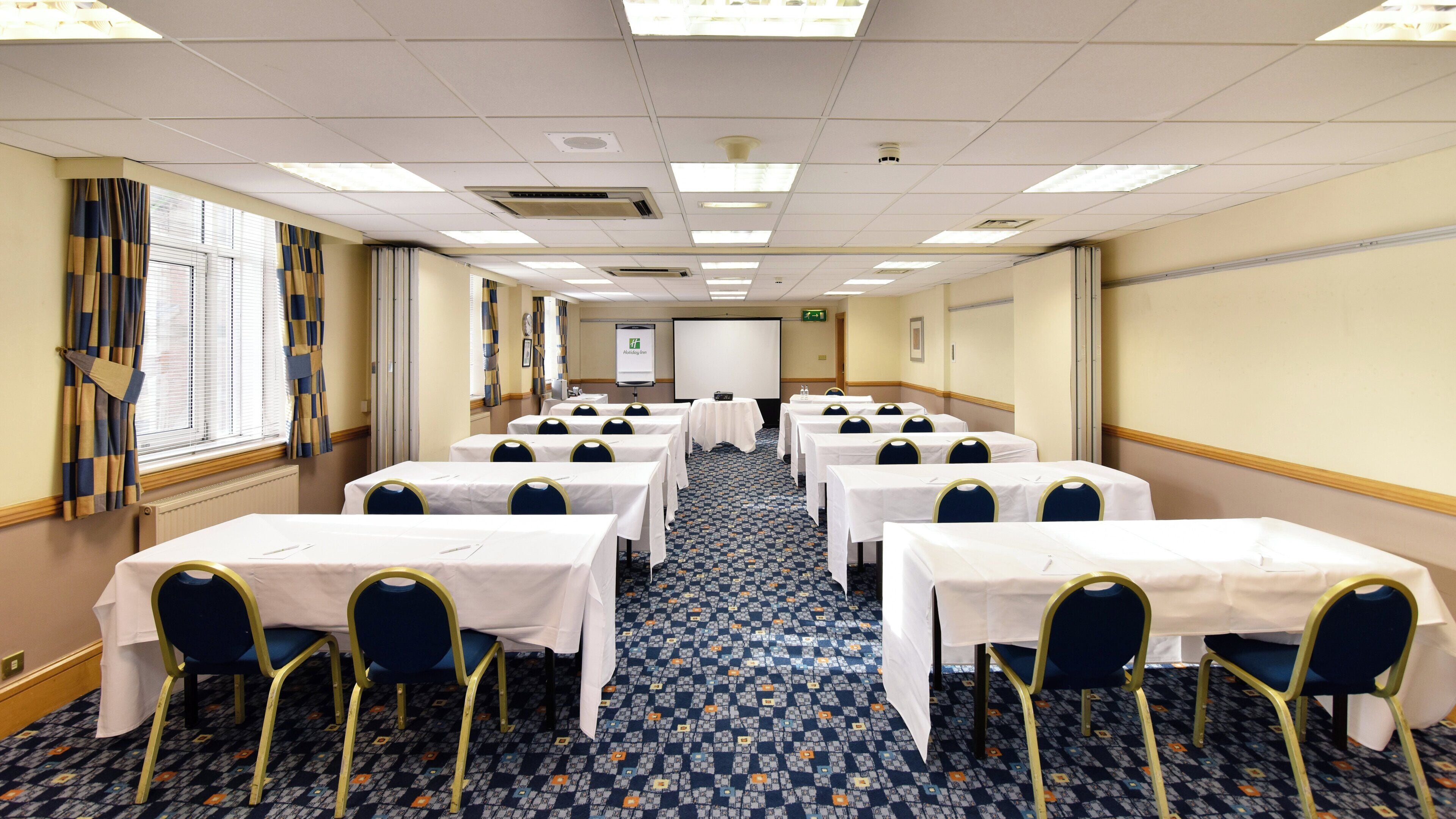 A conference room with rows of tables and chairs, a projector screen, and a carpeted floor.