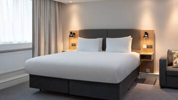 Ein modernes Hotelzimmer mit einem großen Bett, zwei Nachttischen mit Lampen, einem grauen Sessel und Blick ins Freie durch ein Fenster mit Vorhängen.