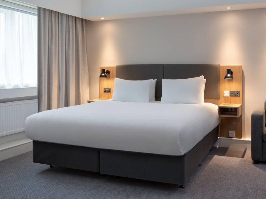 Ein modernes Hotelzimmer mit einem großen Bett, zwei Nachttischen mit Lampen, einem grauen Sessel und Blick ins Freie durch ein Fenster mit Vorhängen.