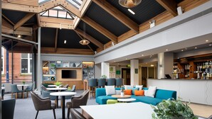 A spacious lounge area with a high ceiling, wooden beams, and a bar with a variety of bottles. There are seating areas with tables and chairs, and a television mounted on the wall.
