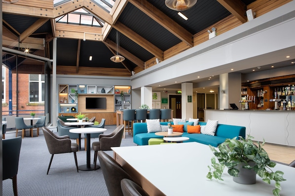 A spacious lounge area with a high ceiling, wooden beams, and a bar with a variety of bottles. There are seating areas with tables and chairs, and a television mounted on the wall.