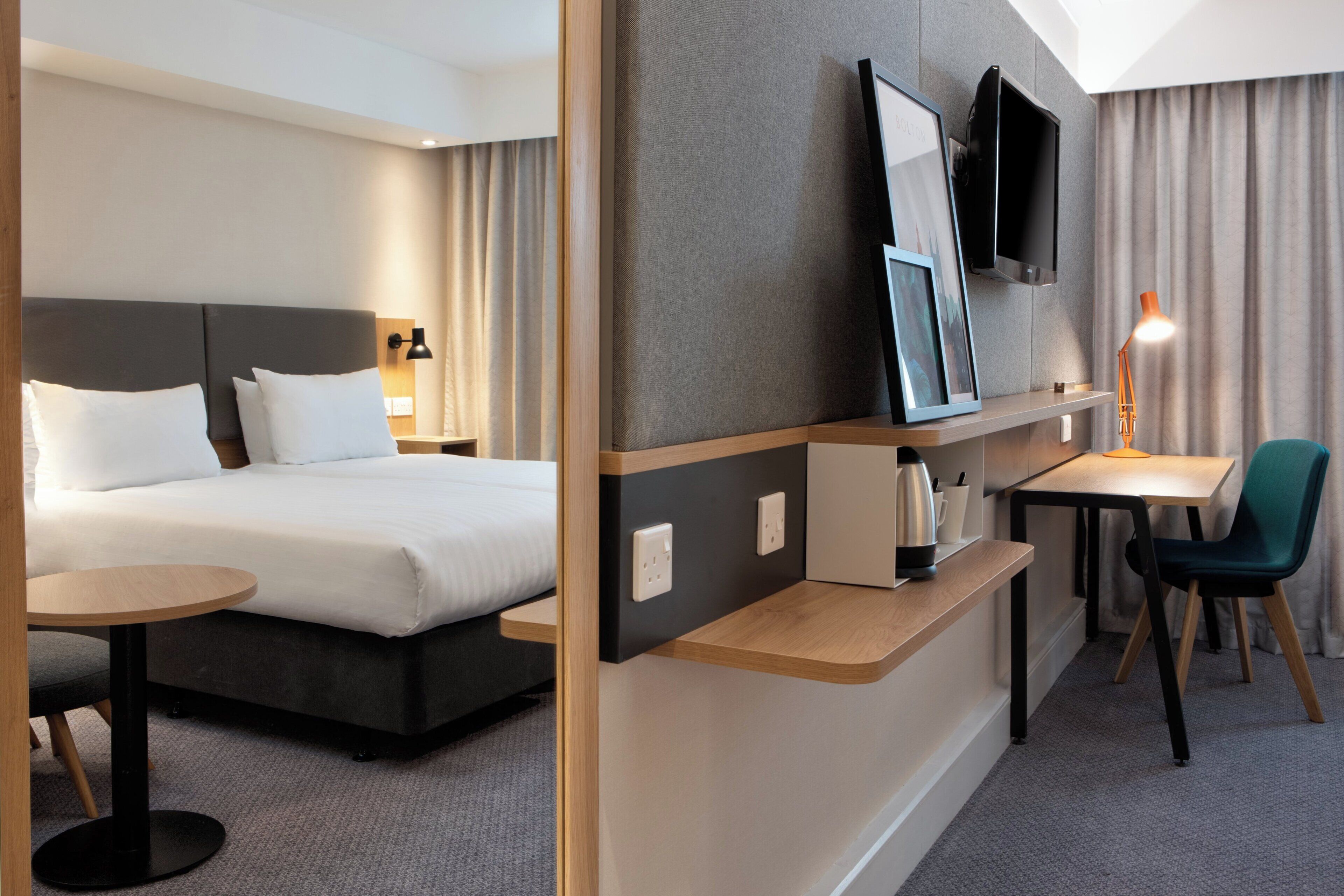 A hotel room with a bed, bedside table, lamp, TV, and a desk with chair.