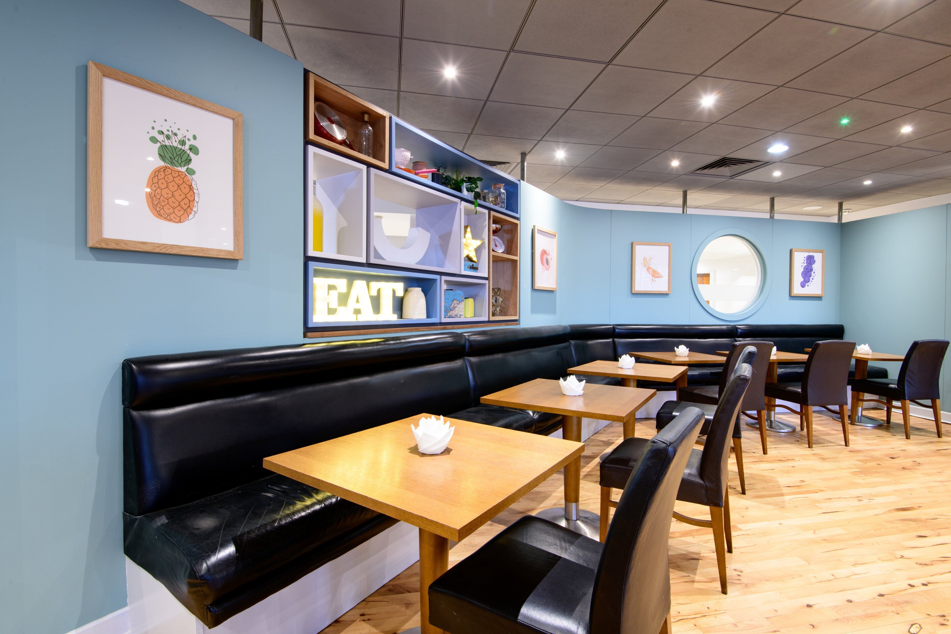A restaurant interior with a black leather booth seating area, wooden tables, and a blue wall with framed artwork.