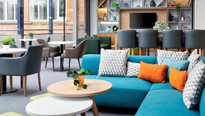 A modern lounge area with a blue sofa, multiple chairs, a round wooden table, and a TV mounted on the wall.