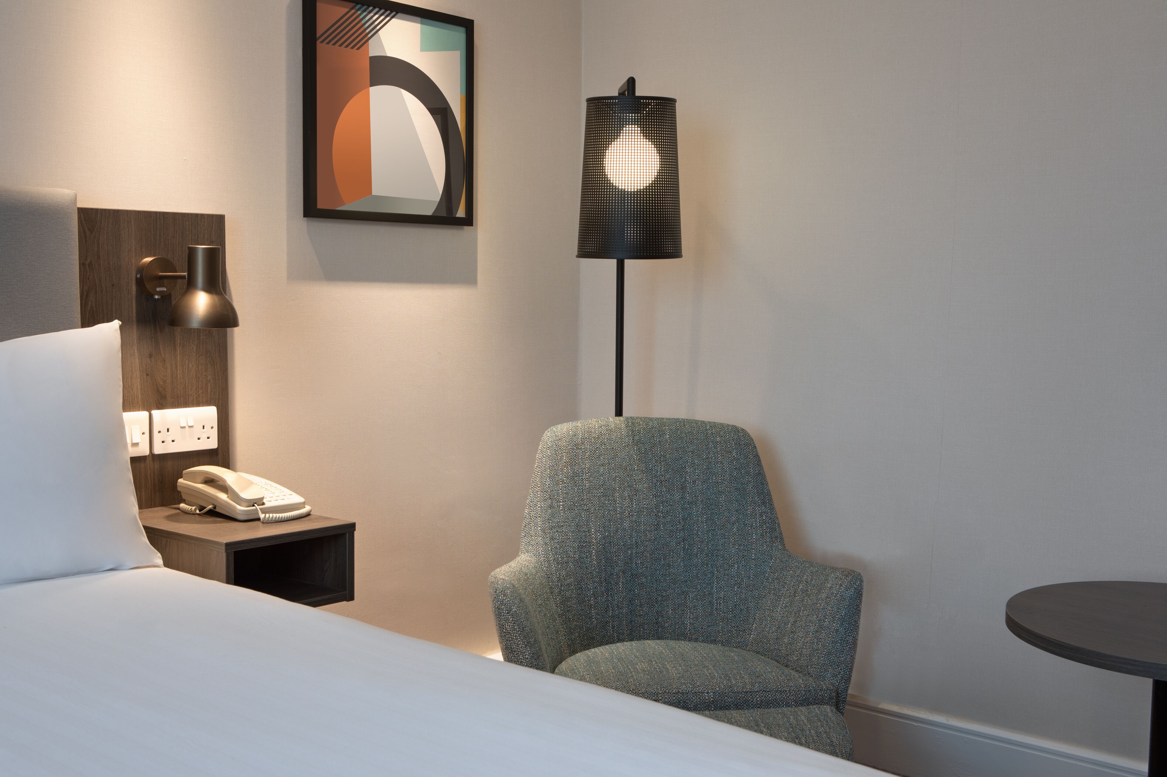 A hotel room with a bed, a chair, a bedside table with a phone, a lamp, and a painting on the wall.
