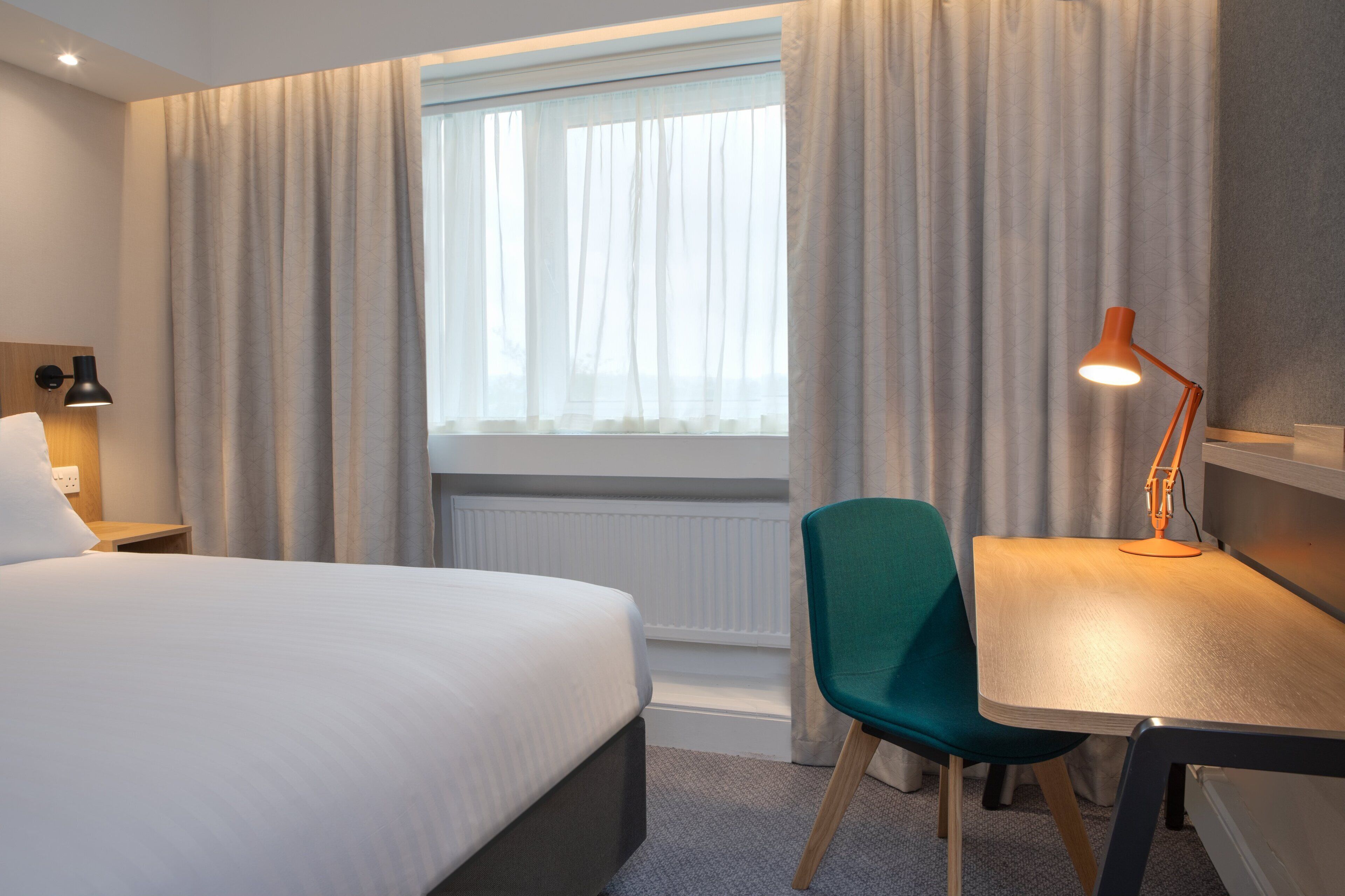 A hotel room with a bed, a desk, a chair, a lamp, and a window with curtains.