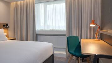A hotel room with a bed, a desk, a chair, a lamp, and a window with curtains.