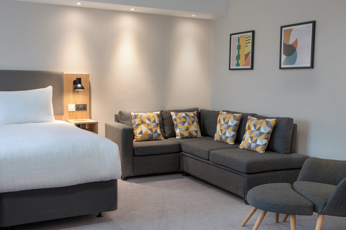 Une chambre d’hôtel moderne avec un lit, un canapé gris agrémenté de coussins à motifs, un fauteuil gris et deux tableaux abstraits accrochés au mur.