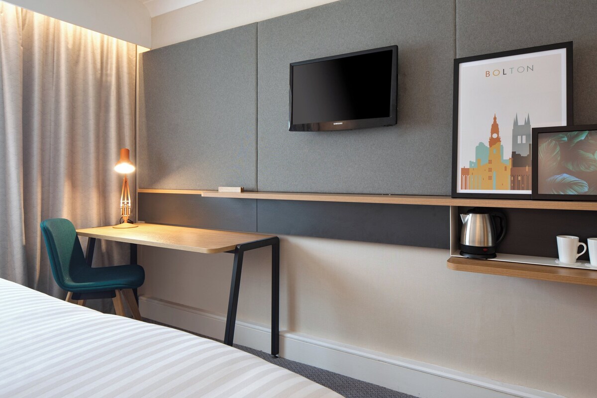 Une chambre d’hôtel comprenant un lit, un bureau avec une chaise, un téléviseur à écran plat, un tableau représentant un paysage urbain, une machine à café et une tasse posée sur une étagère.