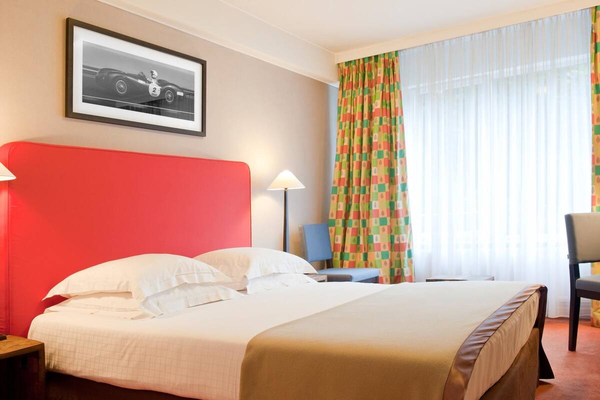 Ein Hotelzimmer mit einem großen Bett, einem roten Kopfteil, einem gerahmten Bild an der Wand, einem bunten Vorhang und einem Sessel.